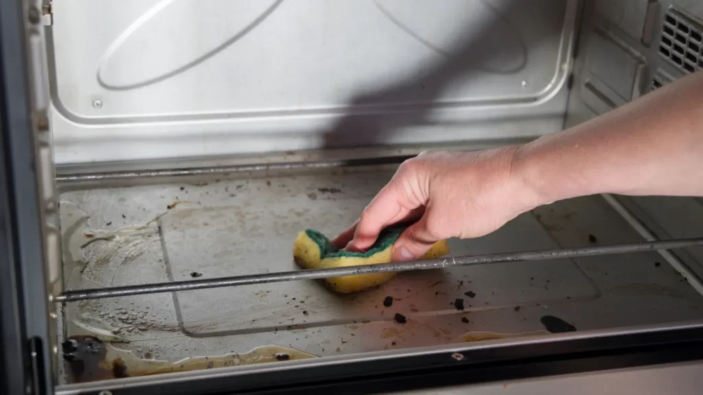 The image displays a close-up of a person's hand cleaning the interior of an oven. The oven has visible stains and food residue on the bottom, and the person is using a yellow and green sponge to scrub the surface. The act of cleaning suggests the removal of baked-on grease and spills, a common maintenance task to keep kitchen appliances in functional and hygienic condition. The oven door is open, allowing access to the interior, and the lighting indicates the oven is turned off during the cleaning process.
